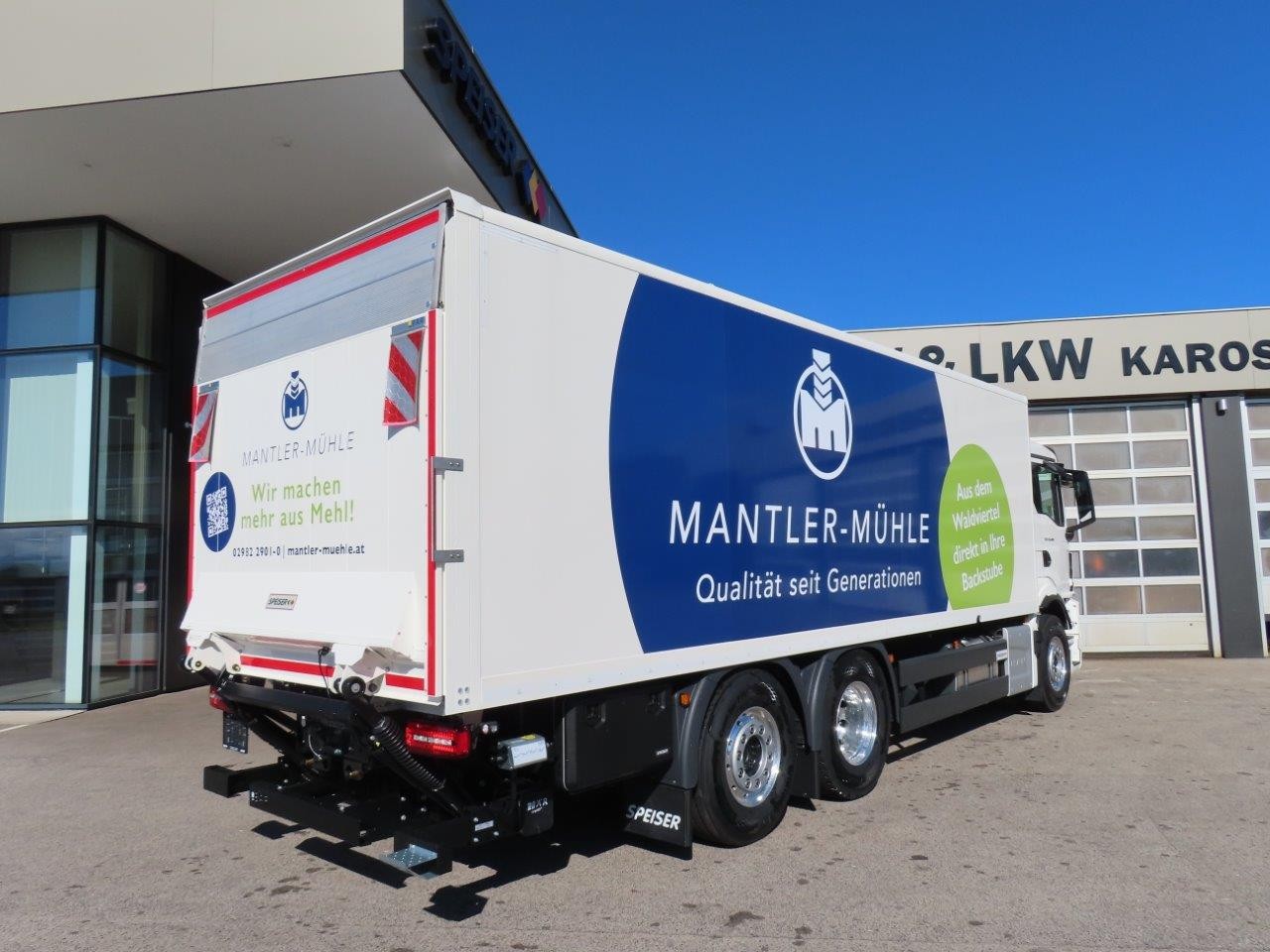 Vor einem Gebäude steht ein großer weißer Lkw mit der Aufschrift „MANTLER-MÜHLE“ und einem blauen Logo. An der Seite des Lkws steht „Qualität seit Generationen“. An der Hecktür befindet sich ein Schild, das auf Mehlprodukte hinweist.