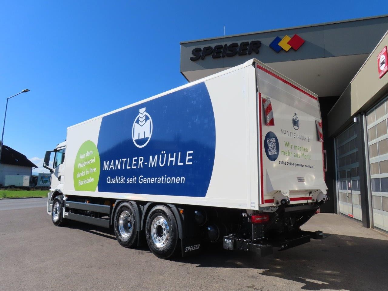Ein weißer Lkw mit dem Mantler-Mühle-Logo steht in der Nähe eines Gebäudes, an dessen Fassade der Name „Speiser“ zu lesen ist. Der Lkw ist an den Seiten und am Heck mit blauen und grünen Werbegrafiken versehen.
