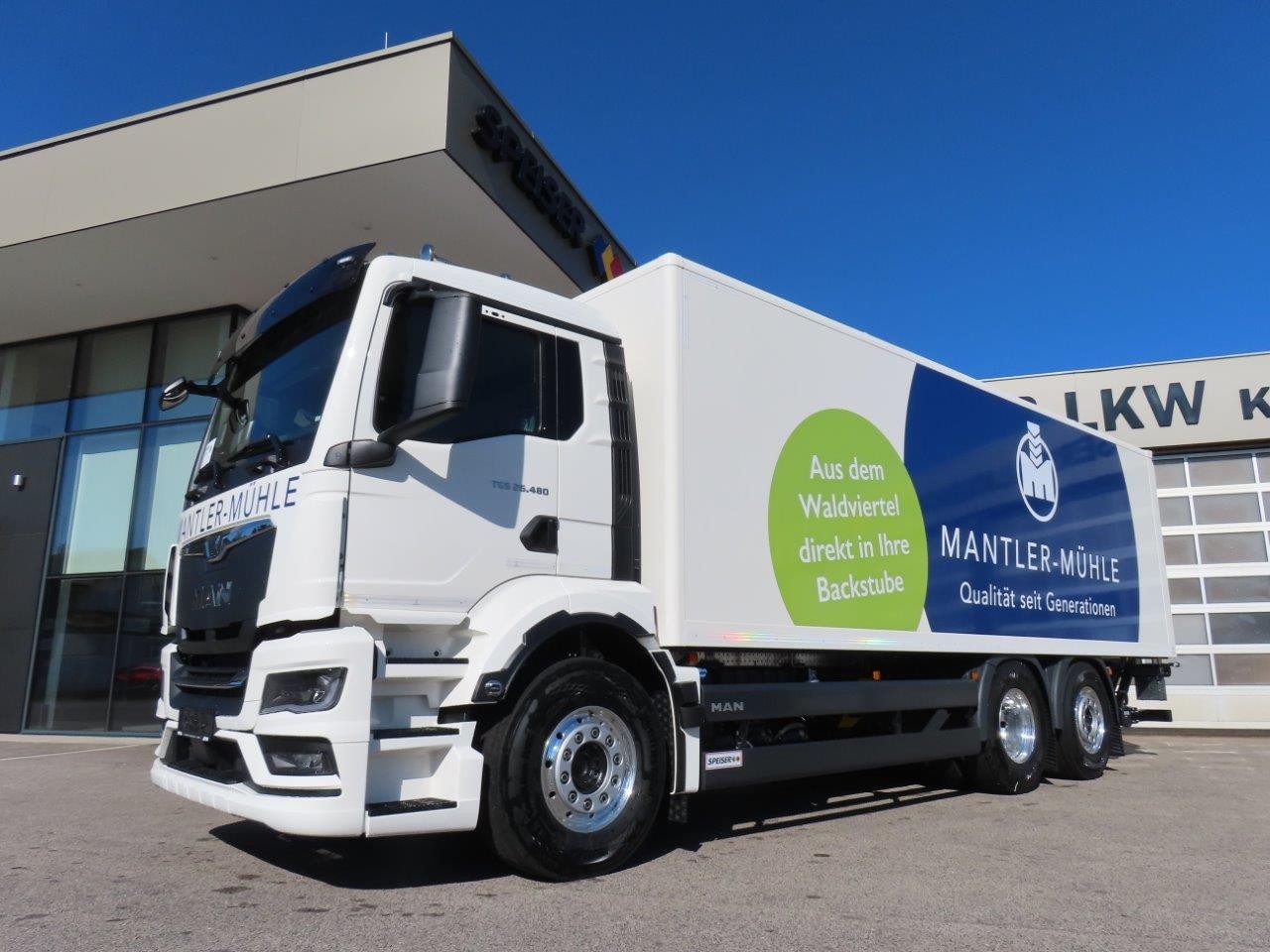 Vor einem modernen Gebäude steht ein weißer MAN-Lieferwagen mit dem Mantler-Mühle-Logo. An der Seite des Lkw sind grüne und blaue Werbegrafiken angebracht.