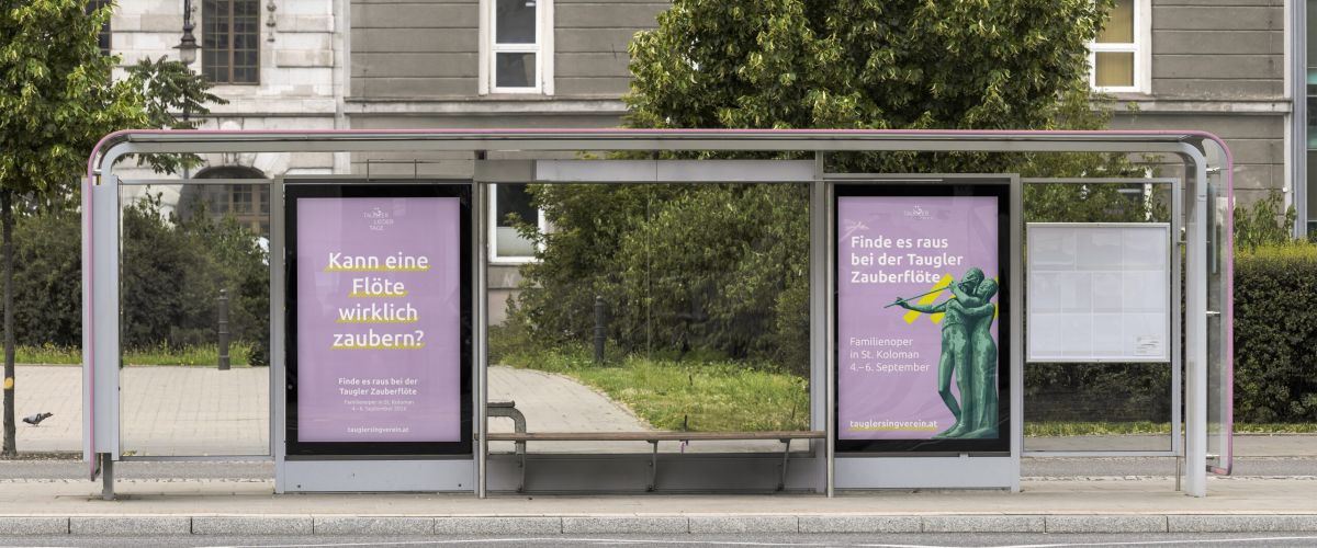 Eine Straßenbahnhaltestelle mit zwei violetten Plakaten auf Deutsch, dahinter Bäume und eine leere Bank im Wartehäuschen. Im Vordergrund sind die Straße und die Straßenbahnschienen zu sehen.