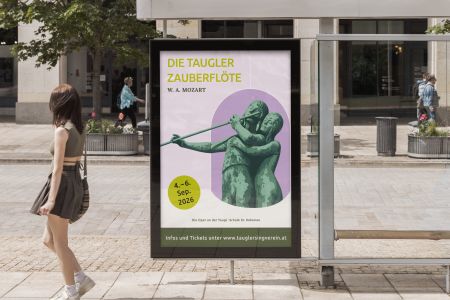 Eine Bushaltestellenanzeige wirbt für „Die Zauberflöte“ von Taugler mit Statuen, die Flöte spielen, den Veranstaltungsterminen und einer Website. Eine Frau geht auf einem städtischen Bürgersteig vorbei, im Hintergrund sind Bäume und Gebäude zu sehen.